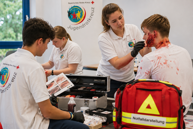 Vier Jugendliche, von denen einer eine Schminkanleitung studiert, eine junge Frau einen Darsteller eine Verletzung im Gesicht schminkt, die vierte Person ist im Hintergrund beschäftigt.